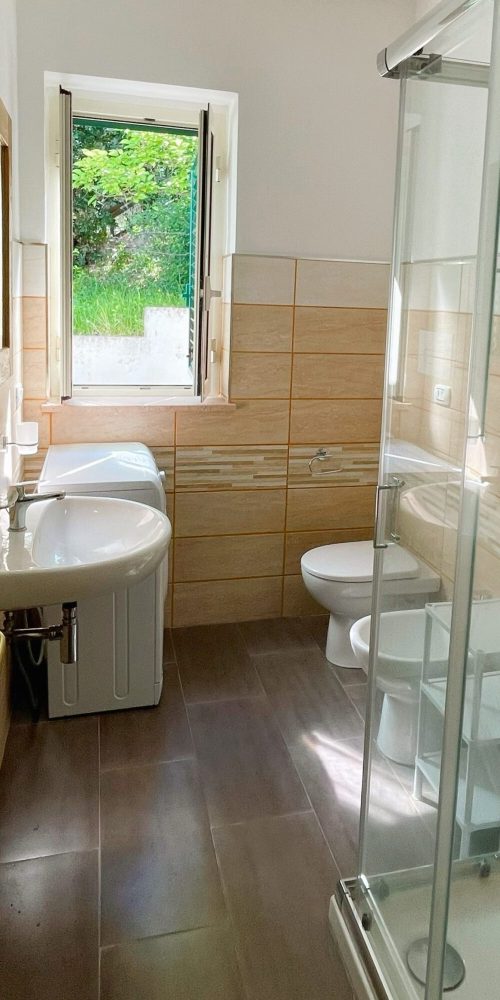 Bagno moderno con doccia, lavabo e finestra aperta su vista verde.